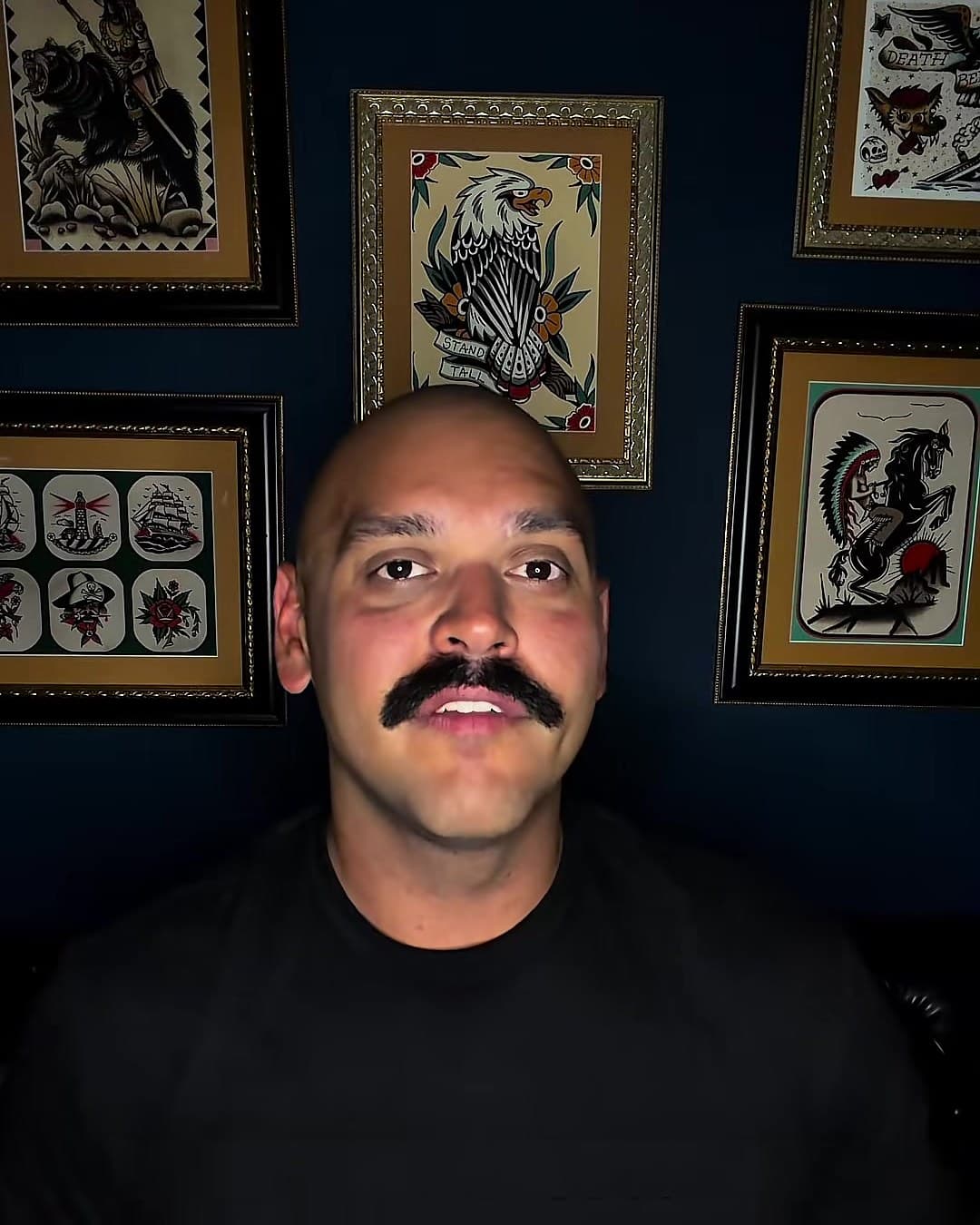 Kody talking to camera in his studio, framed tattoo flash on a navy wall behind him.