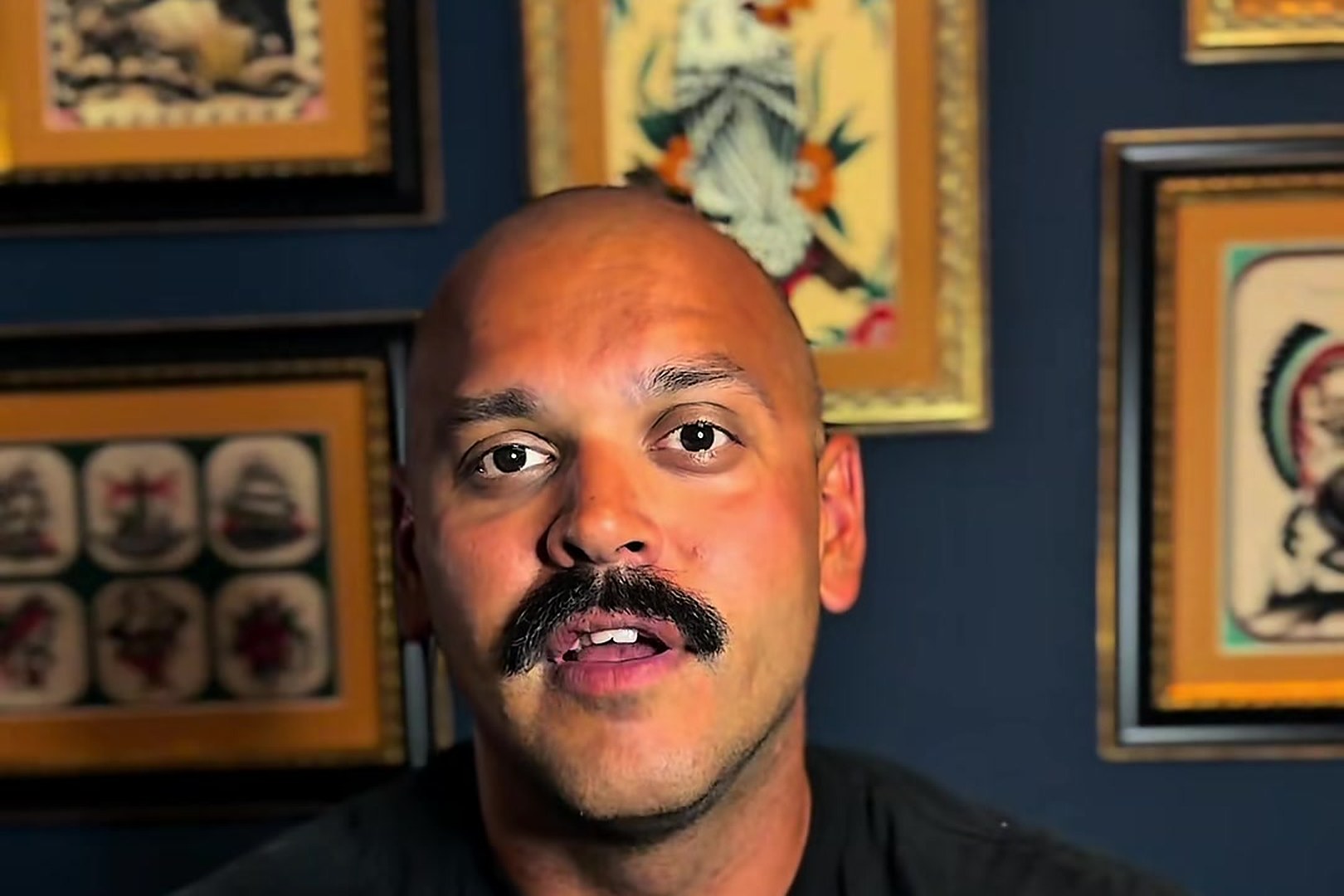 Kody, bald with a thick mustache and a black T-shirt, talking to camera in front of a wall of framed tattoo flash.